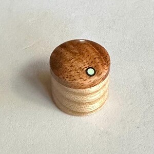 May include: A small wooden box with a light brown and dark brown wood grain pattern. The lid has a small green circle inlay.