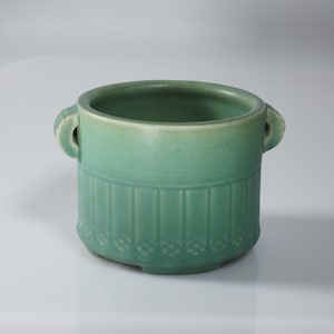 May include: A teal ceramic planter with two small handles. The planter has vertical grooves and a diamond pattern near the base. The interior is a darker shade of teal. The planter is a decorative item.