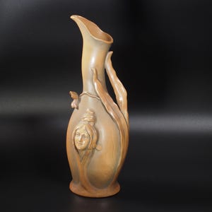 Op de afbeelding: Een hoge, beige decoratieve vaas met een slanke hals en een gebogen handvat. De vaas heeft een gebeeldhouwd vrouwengezicht en bladvormige details. Het ontwerp is elegant en artistiek.