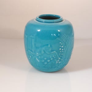Op de afbeelding: Een turquoise keramische vaas met een ronde vorm en een smalle hals. De vaas heeft reliëfontwerpen van herten en andere patronen. De glanzende afwerking reflecteert licht, waardoor de details van het reliëf worden benadrukt. Een decoratief stuk voor woondecoratie.