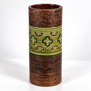 Puede incluir: Un jarrón cilíndrico de cerámica con un exterior de color marrón oscuro texturizado y una banda horizontal ancha en el centro. Esta banda es de color verde claro y está decorada con un patrón repetitivo de motivos florales y líneas curvas.