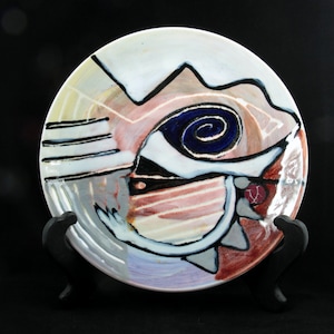 May include: A white ceramic plate with an abstract design featuring a blue eye with a spiral, black lines, and shades of pink, red, and gray.