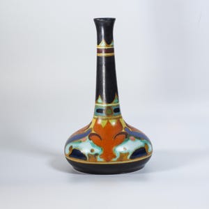 Może przedstawiać: Czarny ceramiczny wazon z długą szyją i pękatą podstawą. Wazon posiada kolorowe wzory kwiatowe i geometryczne w kolorze pomarańczowym, niebieskim, zielonym i żółtym. Wazon ma około 25 cm wysokości.