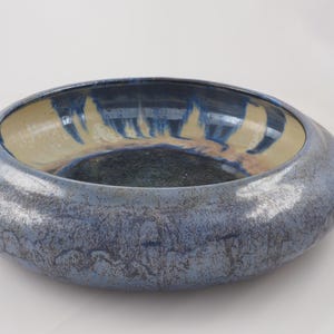May include: A handcrafted ceramic bowl with a wide, shallow design. The exterior has a textured, mottled blue glaze, while the interior features a blend of beige, blue, and brown hues. The bowl is a decorative piece.