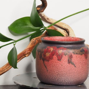 May include: A small, red and brown ceramic pot with a textured surface. The pot has a wide mouth and a rounded body. It is sitting on a silver metal surface.