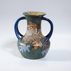 May include: A green ceramic vase with two blue handles and a floral design. The vase features a large, detailed flower in shades of brown and tan, with accents of orange and blue leaves. The rim is a light tan colour.