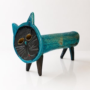 May include: A ceramic cat sculpture featuring a bright turquoise cylindrical body with geometric patterns and four dark brown legs. The circular face is black with two hollow yellow eyes, a small red nose, and etched whiskers, framed by a turquoise border.