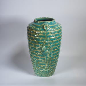 Puede incluir: Un jarrón alto de cerámica turquesa con un patrón de ladrillos texturizados. El jarrón presenta un diseño en relieve de una persona trepando por una cuerda. El jarrón es un objeto decorativo.