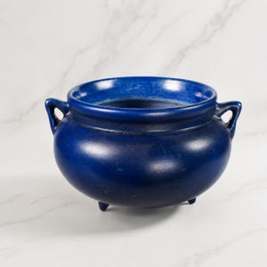 Op de afbeelding: Een diepblauwe keramische kom met twee handvatten en drie kleine voetjes. De kom heeft een afgeronde body en een brede opening. De binnenkant van de kom is een lichtere tint blauw. De kom staat op een wit marmeren oppervlak.