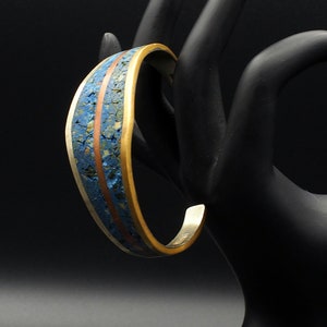 Op de afbeelding: Een zilveren armband met een blauwe en koperen inleg. De armband is op een zwarte displaystandaard.