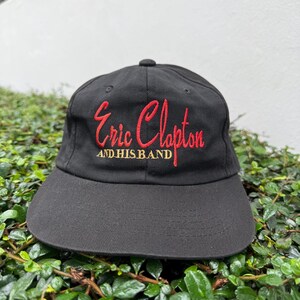 Puede incluir: Gorra de béisbol negra con visera curva. La gorra presenta texto bordado en rojo que dice "Eric Clapton" encima de "AND HIS BAND". La gorra está hecha de una tela duradera y tiene un estilo clásico e informal.