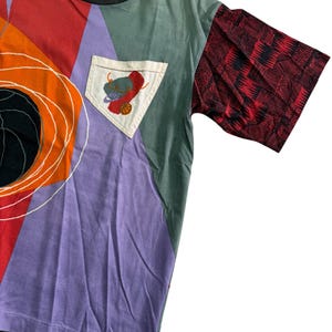 May include: A patchwork t-shirt in vibrant colours. The design includes red, orange, purple, and green panels. One sleeve is red with a black geometric pattern. A pocket features a basketball and a hat. The collar is black.