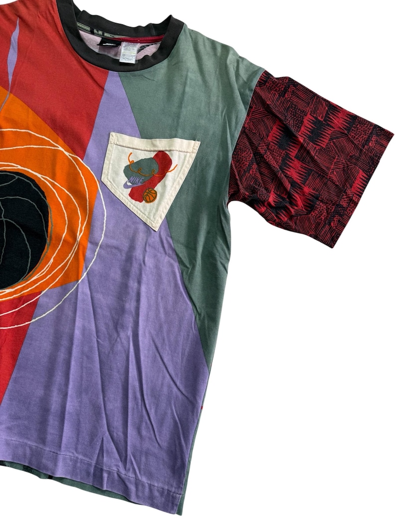 May include: A patchwork t-shirt in vibrant colours. The design includes red, orange, purple, and green panels. One sleeve is red with a black geometric pattern. A pocket features a basketball and a hat. The collar is black.