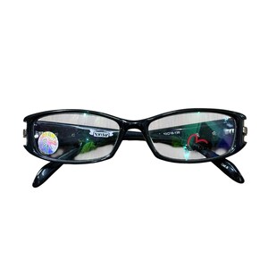 Puede incluir: Gafas con montura negra de forma rectangular. Las gafas tienen un diseño curvo en las patillas y lentes transparentes. La marca "EVISU" es visible en la lente izquierda.