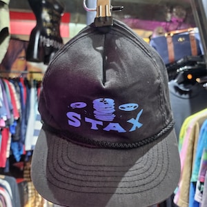 Puede incluir: Gorra de béisbol negra con la palabra "STAX" en azul y morado, junto con un gráfico de discos apilados. La gorra tiene una visera curva y un detalle de cuerda negra en el borde.