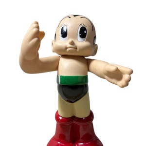 Puede incluir: Una figura vintage de Astro Boy. La figura tiene un cuerpo bronceado, cabello negro y ojos grandes. Viste pantalones cortos negros, un cinturón verde y botas rojas. Los brazos están extendidos y la figura posa con una mano levantada.