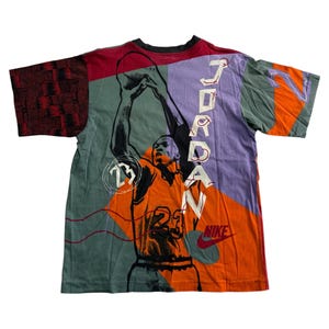 May include: A vibrant t-shirt with a basketball player graphic, the name "JORDAN" printed vertically on the back. The shirt has a patchwork design with orange, purple, green, and red panels, and the Nike logo.