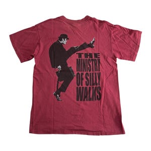May include: A maroon t-shirt featuring a black and white graphic of a man in a suit and hat, mid-stride, with the text "THE MINISTRY OF SILLY WALKS" below. The shirt has short sleeves and a crew neck.
