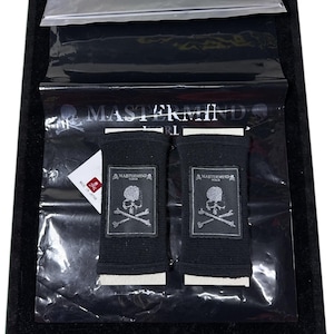 May include: Two black wristbands with a skull and crossbones logo, displayed in a clear plastic bag. The wristbands are on a black velvet surface. The logo is silver and the text "MASTERMIND" is visible.