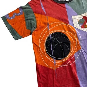 May include: A multi-coloured t-shirt with a basketball design. The shirt has a black basketball graphic with white circular lines on a red and orange background. The shirt has a pocket with a basketball graphic and the Nike logo.