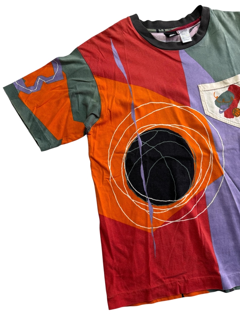May include: A multi-coloured t-shirt with a basketball design. The shirt has a black basketball graphic with white circular lines on a red and orange background. The shirt has a pocket with a basketball graphic and the Nike logo.