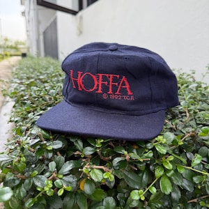 Puede incluir: Gorra de béisbol azul marino con la palabra "HOFFA" bordada en rojo en la parte delantera. El texto "© 1992 T.C.R." está bordado en rojo debajo. La gorra está sobre un arbusto verde.