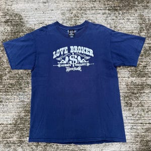 Puede incluir: Camiseta azul marino con la inscripción blanca "LOVE BROKER", con un diseño de signo de dólar y alas. Las palabras "ALWAYS QUALITY" y "Third Rail" también están impresas en la camiseta.