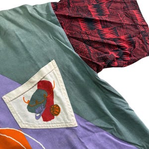 May include: A patchwork long-sleeved shirt with a unique design. The shirt features panels of green, purple, and orange fabric. The right sleeve is red with a black geometric pattern. A white pocket displays a Nike logo with a basketball.