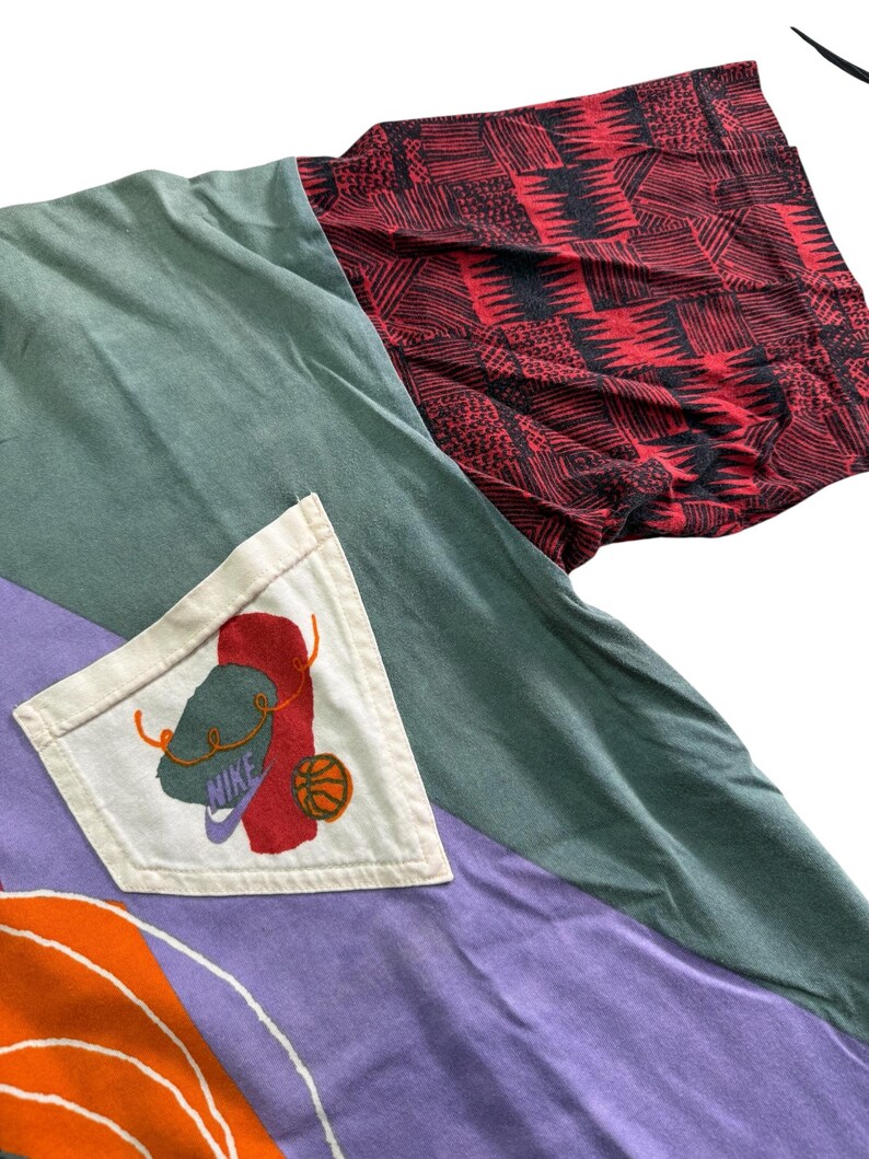 May include: A patchwork long-sleeved shirt with a unique design. The shirt features panels of green, purple, and orange fabric. The right sleeve is red with a black geometric pattern. A white pocket displays a Nike logo with a basketball.