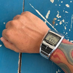 May include: A silver-toned Casio digital watch with a rectangular face and a black display panel. The screen shows the time as 9:47 and includes the text 'BETA REZ' and 'LIGHT' on the lower black buttons. The watch features a metallic finish.