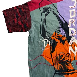 May include: A vintage t-shirt with a basketball player graphic in orange and black, with the number 23. The shirt has a colour block design with red, green, and purple panels. The sleeve features a red and black geometric pattern.