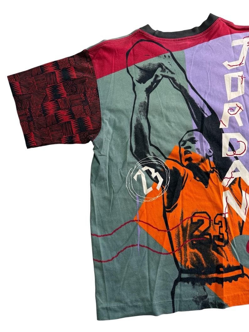 May include: A vintage t-shirt with a basketball player graphic in orange and black, with the number 23. The shirt has a colour block design with red, green, and purple panels. The sleeve features a red and black geometric pattern.