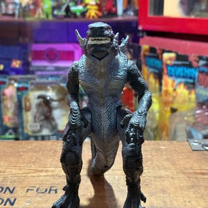 Può includere: Action figure grigia e nera di una creatura dall'aspetto rettiliano. La figura ha la pelle strutturata, denti affilati ed è in posa a quattro zampe. Giocattolo da collezione.