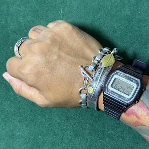 Puede incluir: Se muestra un anillo de plata, una pulsera de plata con diseño de pluma y un reloj digital negro con esfera plateada. El reloj muestra la hora 6:24:25. El fondo es de color verde liso.