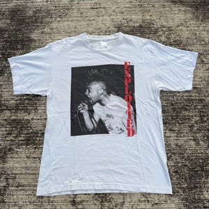 Pode incluir: Camiseta branca com uma imagem em preto e branco de uma pessoa cantando em um microfone. A palavra "EXPLOITED" é impressa verticalmente em vermelho no lado direito da imagem. A camisa é feita de um material macio e leve.