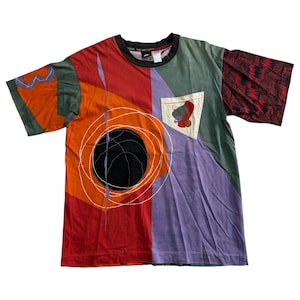 May include: A vibrant, short-sleeved t-shirt with an abstract design. The shirt has panels of red, orange, purple, and green. A black circle with white lines and a pocket with a graphic are visible. The collar is black.