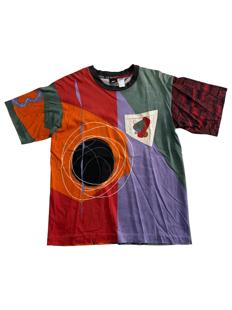 May include: A vibrant, short-sleeved t-shirt with an abstract design. The shirt has panels of red, orange, purple, and green. A black circle with white lines and a pocket with a graphic are visible. The collar is black.