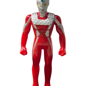 Puede incluir: Figura de acción roja y plateada de un superhéroe con casco plateado y ojos amarillos. La figura tiene un cuerpo rojo con detalles plateados en el pecho y los hombros. La figura está de pie con los puños cerrados.