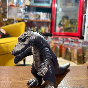 Puede incluir: Figura de dinosaurio gris oscuro y plateado con un diseño texturizado y detallado. El juguete tiene una cola larga, brazos pequeños y una cabeza detallada. La figura está colocada sobre una superficie de madera.