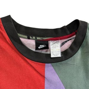 May include: A multi-coloured Nike t-shirt with a black collar. The shirt features a colour block design with red, purple, and green panels. A black Nike logo is visible on the shirt.