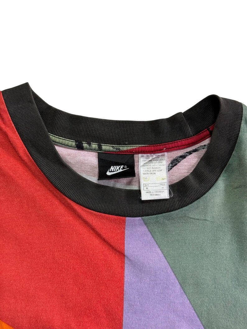 May include: A multi-coloured Nike t-shirt with a black collar. The shirt features a colour block design with red, purple, and green panels. A black Nike logo is visible on the shirt.