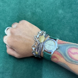 Op de afbeelding: Een pols met een zilveren horloge met een groene leren band en verschillende zilveren armbanden. De armbanden hebben bedels, waaronder een veer en een blad. Een zilveren ring is ook zichtbaar.