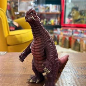 Può includere: Una statuetta di dinosauro color marrone con un corpo squamoso e strutturato. Il giocattolo ha una testa dettagliata con denti e artigli visibili. La statuetta è in piedi su una superficie di legno.