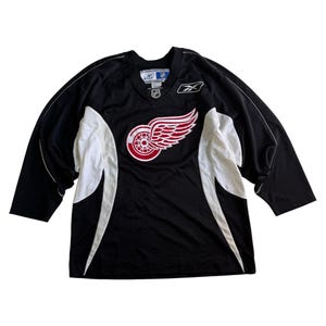 May include: Black hockey jersey with white accents and the Detroit Red Wings logo on the front. The logo features a red wheel with white wings. The jersey has a V-neck and long sleeves. Reebok logo on the right chest.