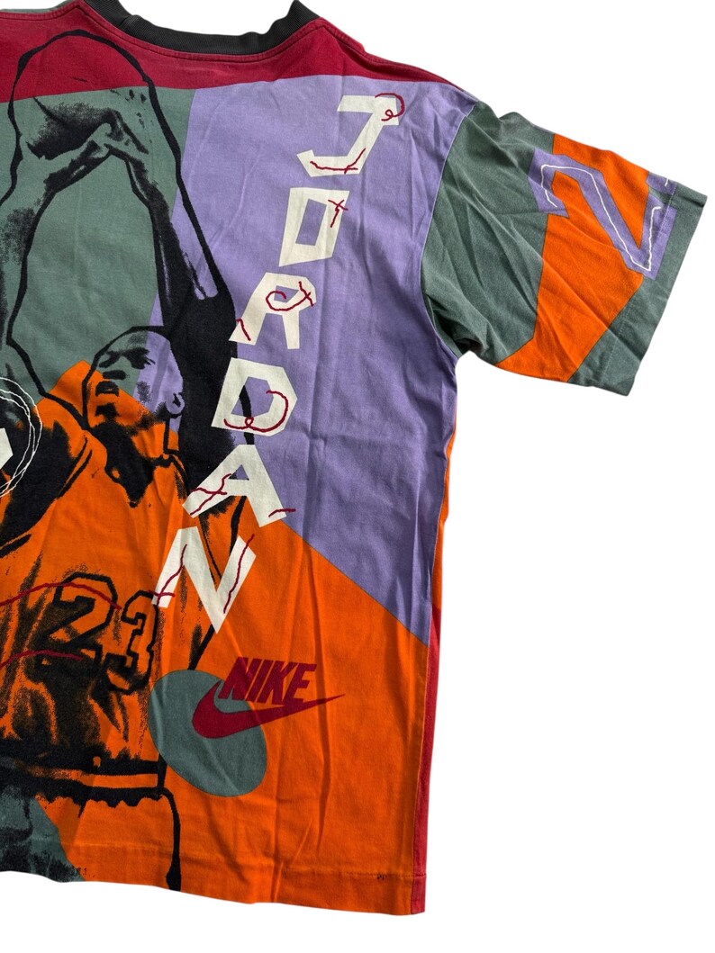 May include: A vintage graphic t-shirt in orange, purple, and green, featuring a basketball player and the word "JORDAN" in white. The Nike logo is visible. The shirt has a crew neck and short sleeves.
