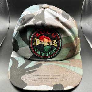 Könnte beinhalten: Eine Baseballkappe im Tarnmuster mit einem grünen, roten und gelben gestickten Aufnäher mit der Aufschrift "Zulu Nation Jungle Brothers".