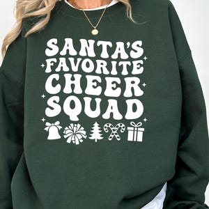 May include: A dark green sweatshirt with the text "SANTA'S FAVORITE CHEER SQUAD" in white, retro-style lettering. Below the text are white graphic icons of a bell, pom-pom, Christmas tree, candy canes, and a gift.