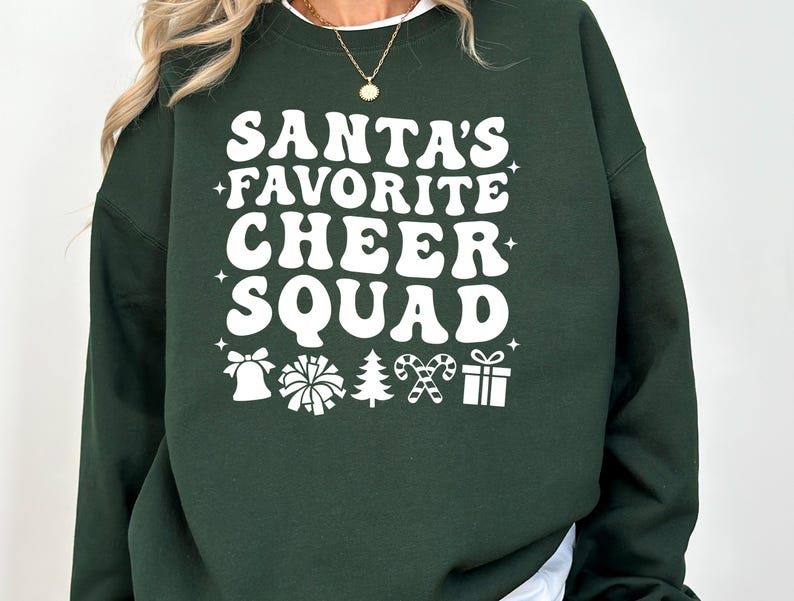 May include: A dark green sweatshirt with the text "SANTA'S FAVORITE CHEER SQUAD" in white, retro-style lettering. Below the text are white graphic icons of a bell, pom-pom, Christmas tree, candy canes, and a gift.
