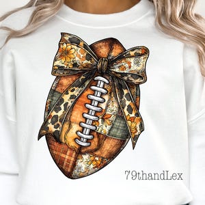May include: A white sweatshirt featuring a graphic of a football decorated with a patchwork pattern of orange, brown, and green plaid and floral prints. A large leopard print bow with orange flowers sits on top. The text 79thandLex is printed below.