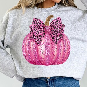 May include: A light gray sweatshirt featuring a large, pink glitter pumpkin with a leopard print bow. The pumpkin has a stem and is covered in pink glitter. The bow is black and brown.